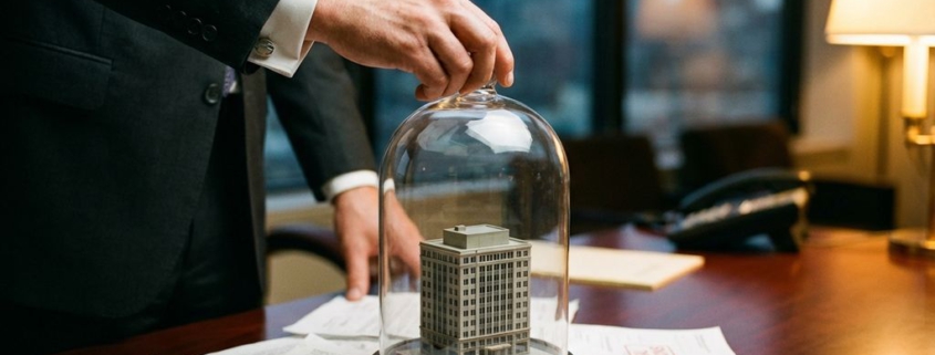 Zakenman in pak plaatst glazen koepel over miniatuur kantoorgebouw op bureau met juridische documenten