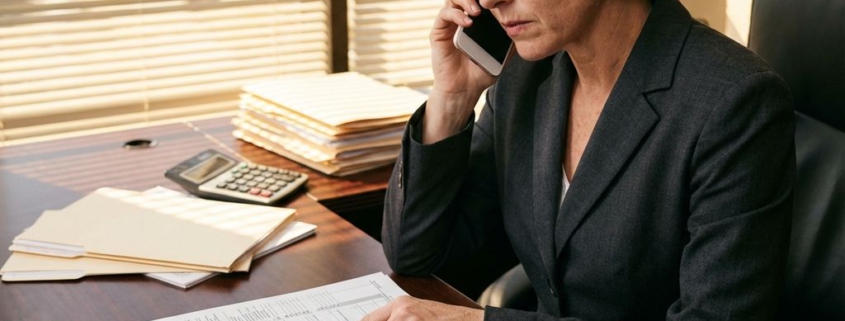 Zakenvrouw in donker pak bekijkt belastingdocumenten aan bureau terwijl ze telefoneert, rekenmachine en facturen zichtbaar