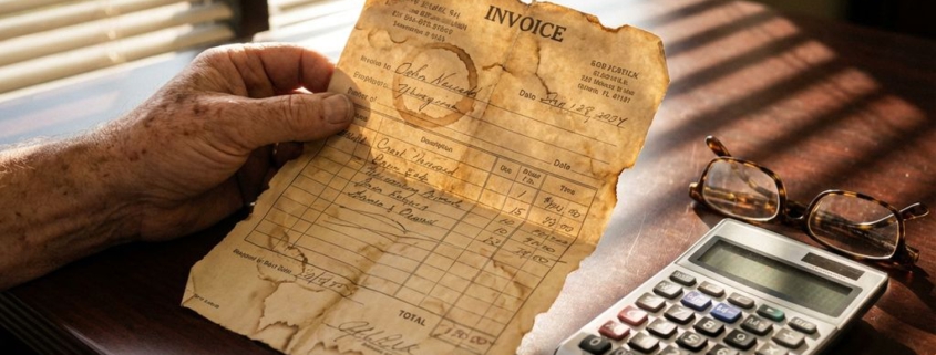 Versleten zakenmannenhand houdt vergeelde factuur vast op mahonie bureau naast calculator en leesbril