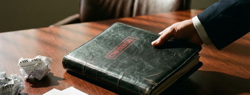 Zakenhand houdt zwart leren map met rode stempel aan mahonie vergadertafel met contracten en vulpen