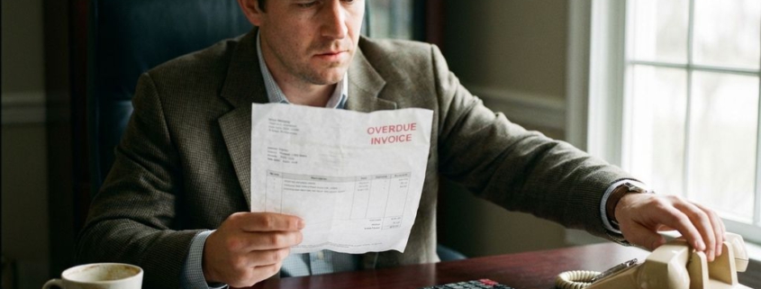 Bezorgde zakenman aan bureau met onbetaalde factuur en telefoon, stapel rekeningen en calculator zichtbaar