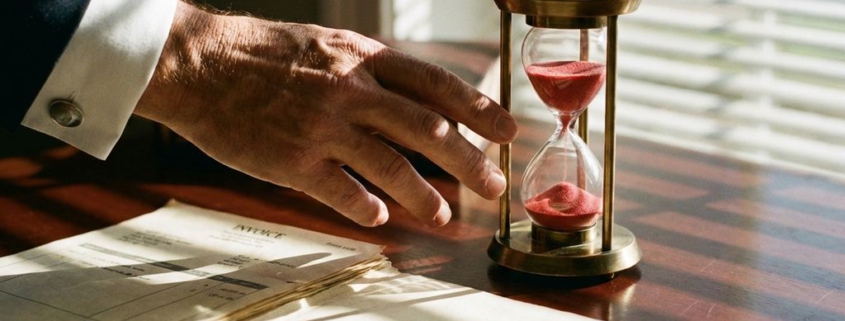 Zakenman reikt naar bijna lege zandloper op bureau met factuurmap en documenten in gouden zonlicht