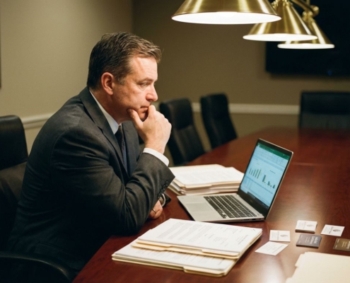 Zakenman in pak zit aan vergadertafel met contracten, documenten van incassobureaus en laptop met financiële spreadsheets