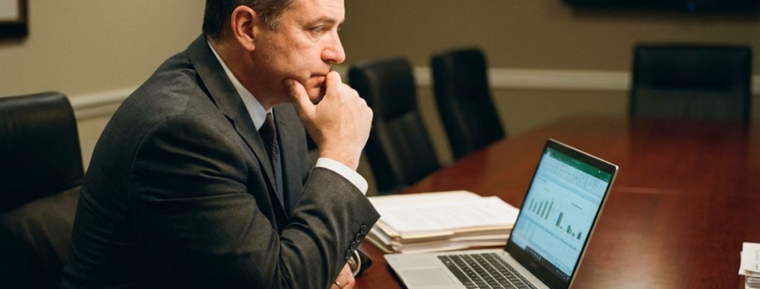 Zakenman in pak zit aan vergadertafel met contracten, documenten van incassobureaus en laptop met financiële spreadsheets