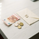 Verfrommeld eurobiljet en losse munten naast een officiële envelop op een wit bureau, bovenaanzicht.
