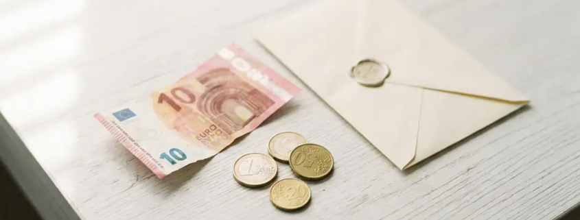 Verfrommeld eurobiljet en losse munten naast een officiële envelop op een wit bureau, bovenaanzicht.