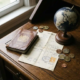 Open versleten leren paspoort op donker eiken bureau naast gevouwen factuur en kleine globe, omringd door buitenlandse munten.