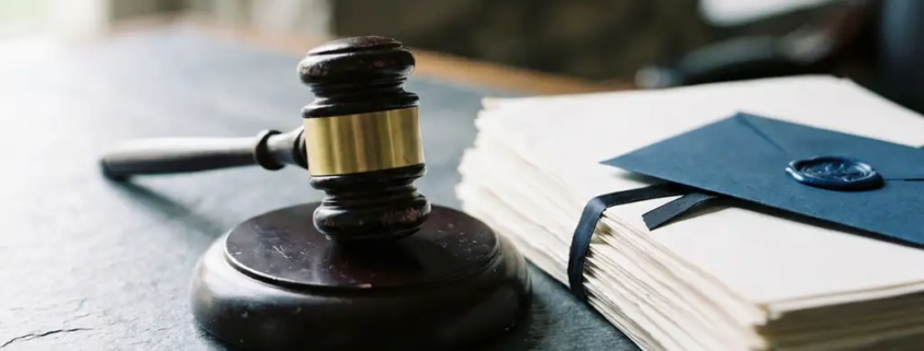 Houten rechtershamer naast gestapelde juridische documenten en verzegelde envelop op stenen bureau, koel grijs en marineblauw licht.