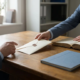 Zakelijke professional schuift betalingsenvelop over houten bureau naar klant, met factuurmap en kasboek op de voorgrond.
